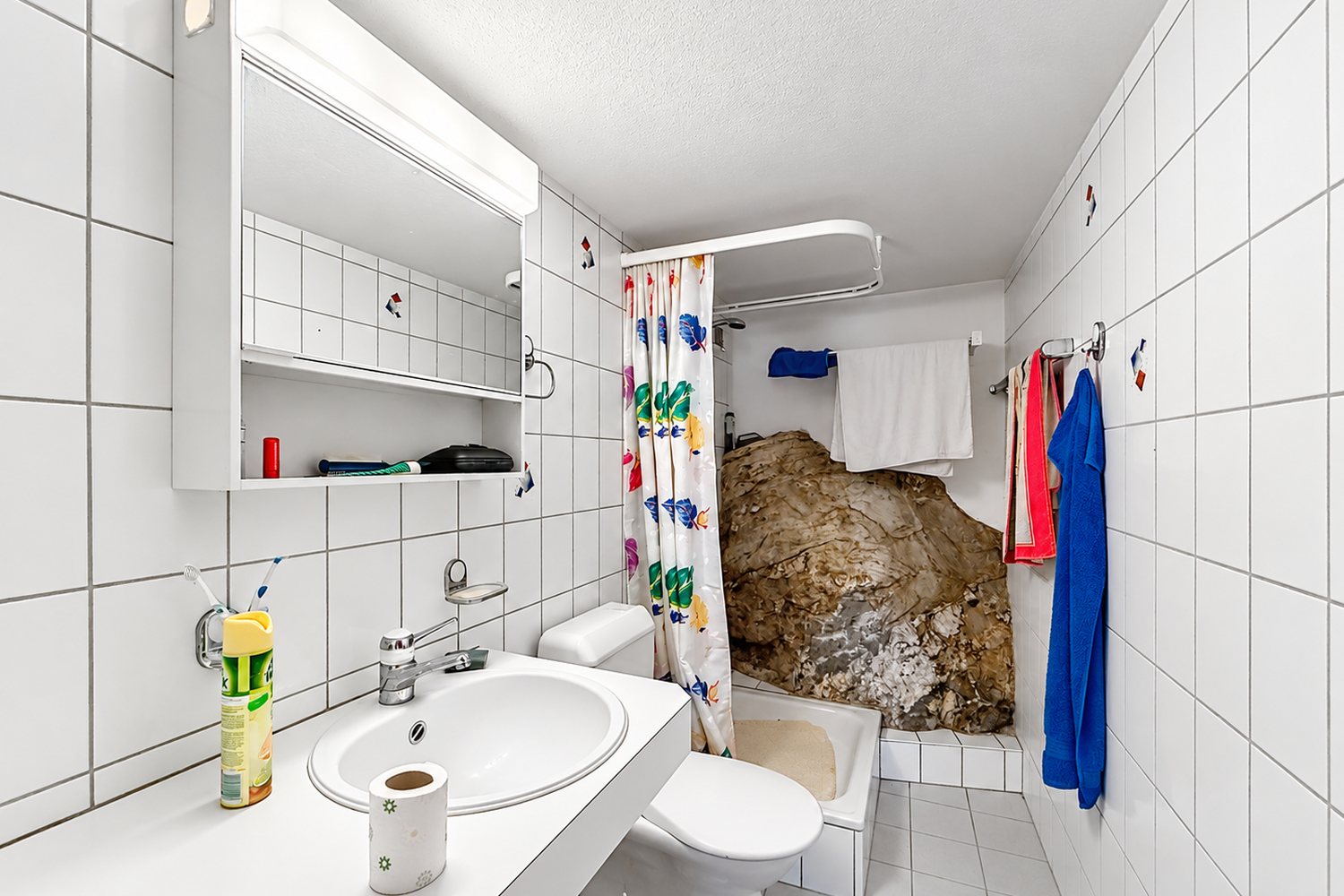 Salle de bains carrelée de blanc avec lavabo, WC, baignoire-douche — et le rocher du Sapey laissé apparent.