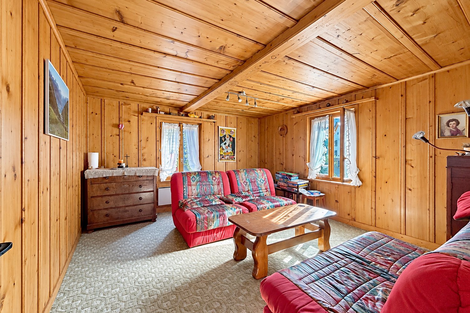 Salon boisé de la maison : lambris et plafond en sapin, canapés, deux fenêtres sur le hameau.