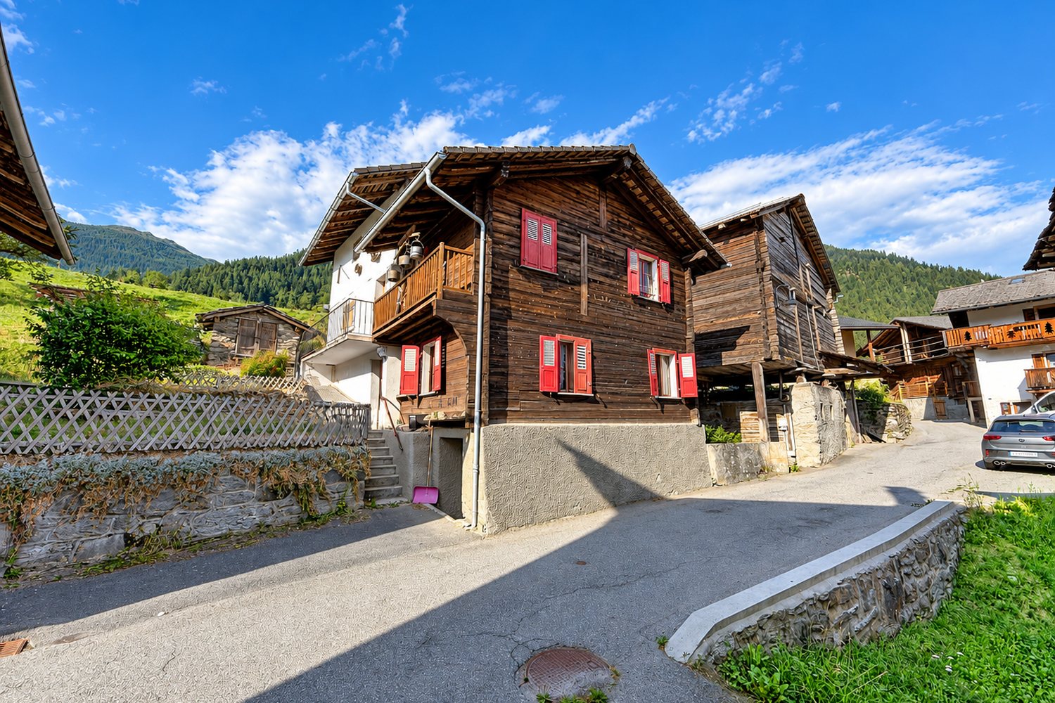La maison dans le hameau du Sapey : madriers patinés, volets rouges, raccard voisin sur pilotis, montagnes en arrière-plan.